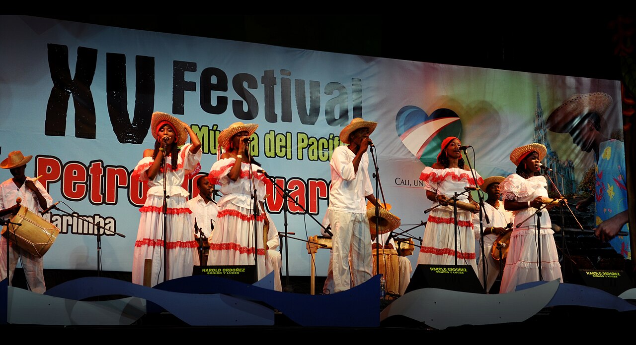 Mujeres y hombres cantan y tocan percusión bajo una pancarta del Festival Petronio Álvarez.