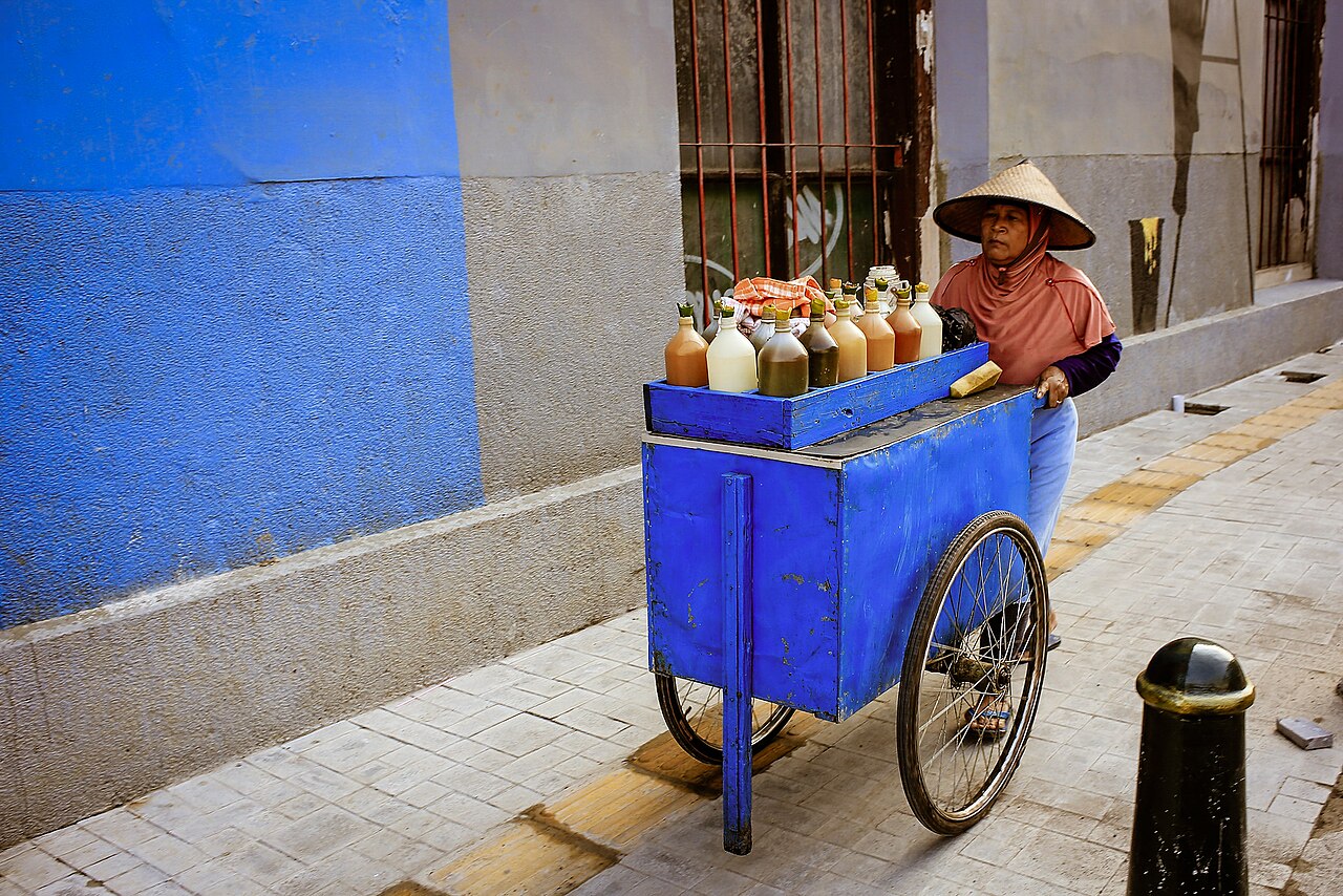 Una mujer con sombrero cónico empuja por una calle pavimentada un carrito azul con botellas de jamu.