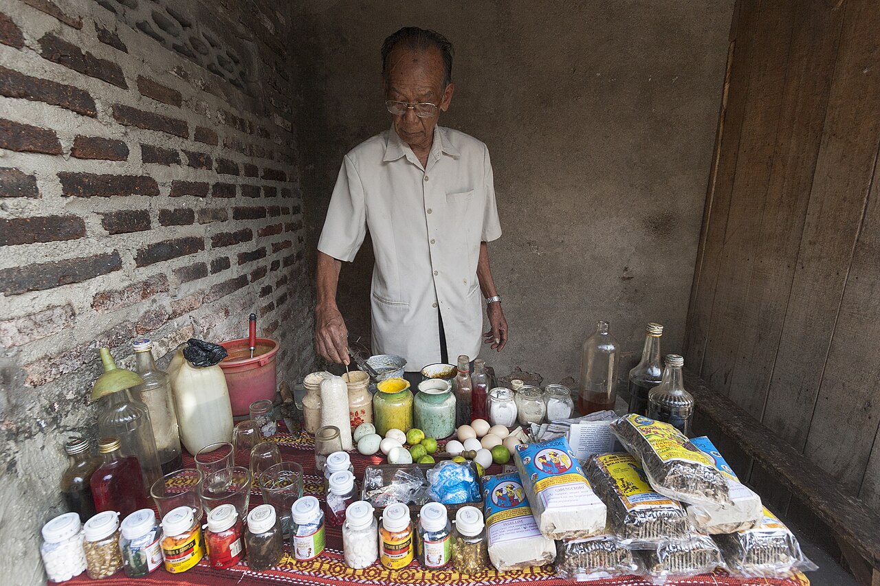 A vendor stands behind bottles, jars and packets at a jamu stall.