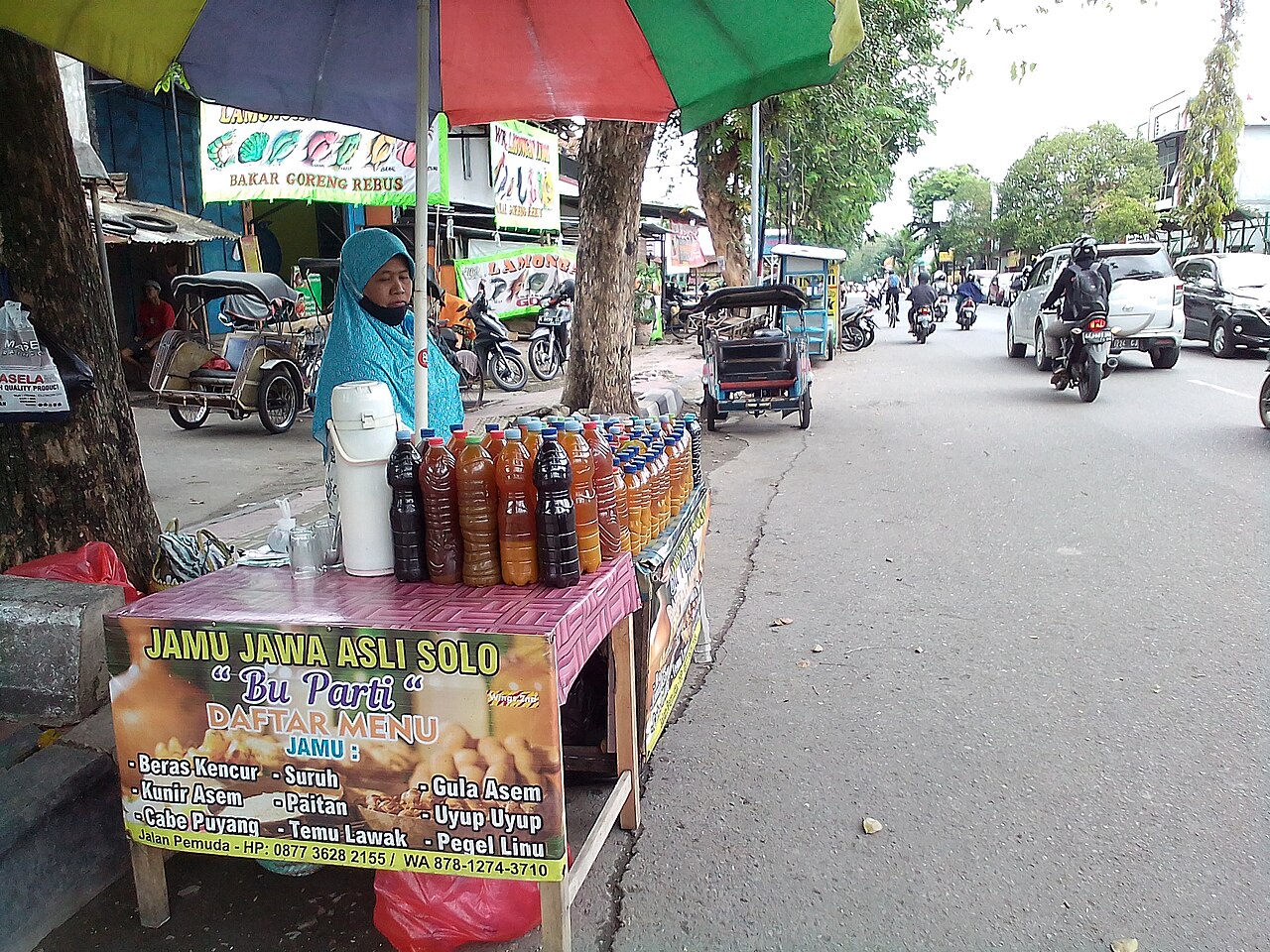 Una mujer detrás de botellas de jamu bajo una sombrilla junto al tráfico.