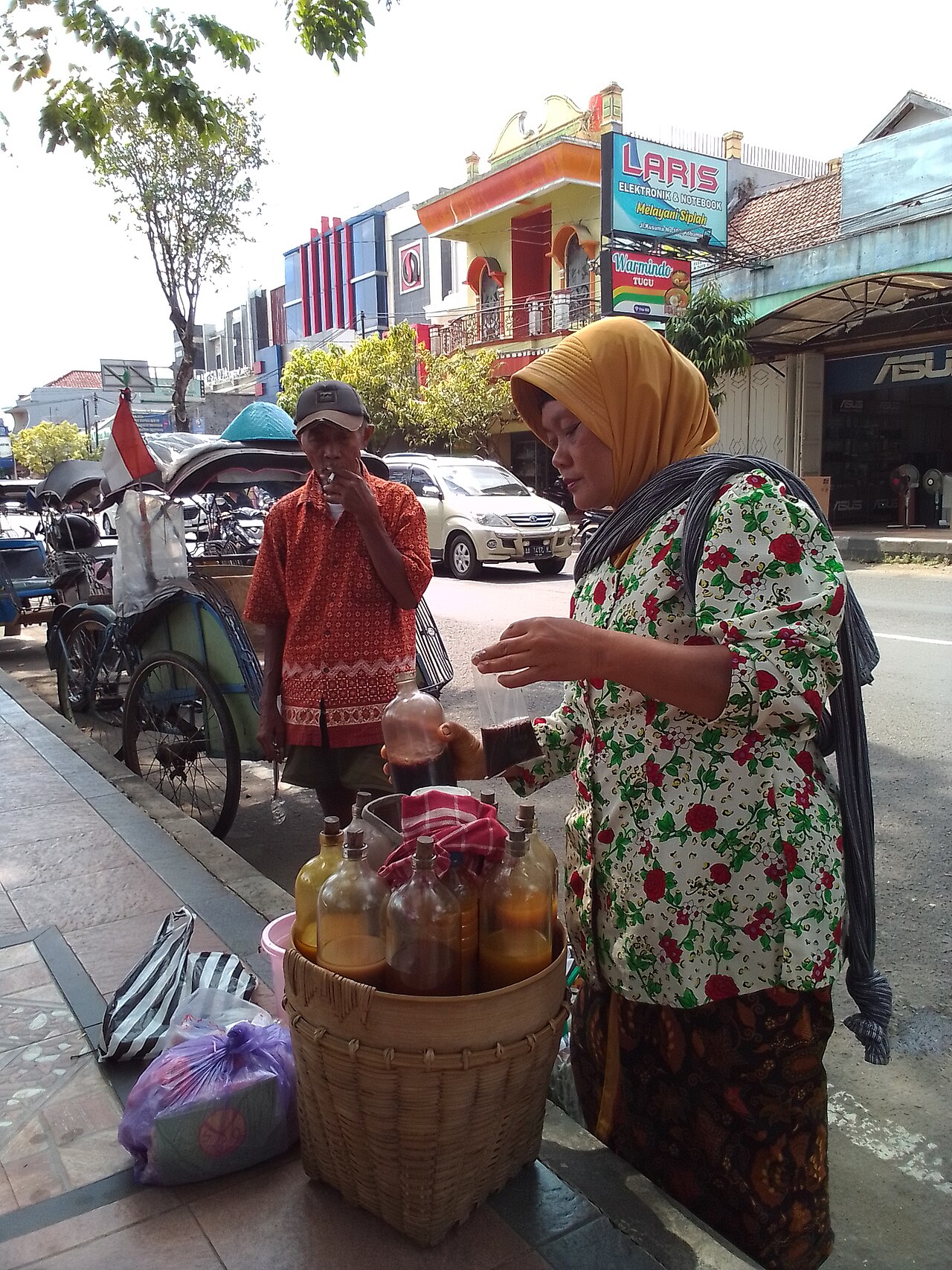 Una mujer sirve jamu junto a unas botellas mientras un hombre espera cerca de un bicitaxi.
