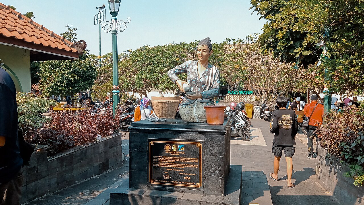 Estatua de una vendedora arrodillada que sirve una bebida, con peatones y motocicletas detrás.