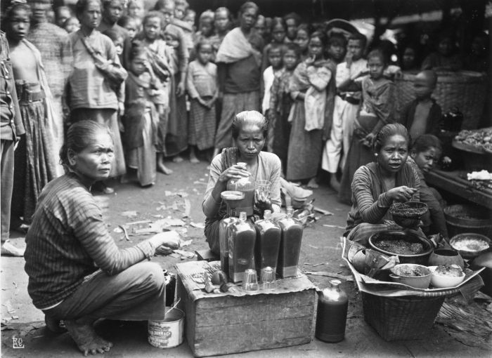 Three seated women prepare jamu among bottles and bowls, with a crowd behind them.