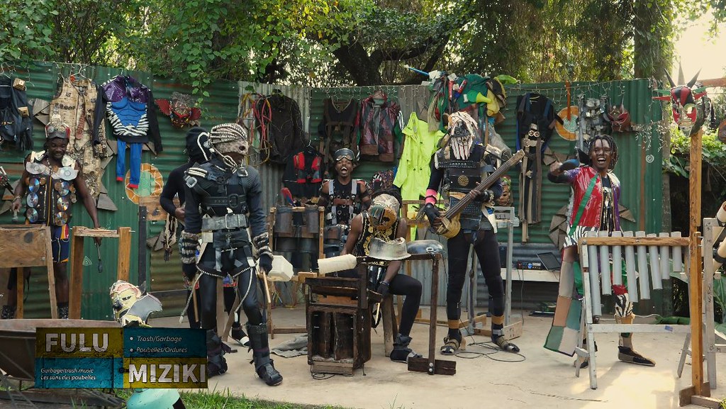 Musicians perform together outdoors, some masked and others with uncovered faces, with tube instruments and hanging costumes around them.