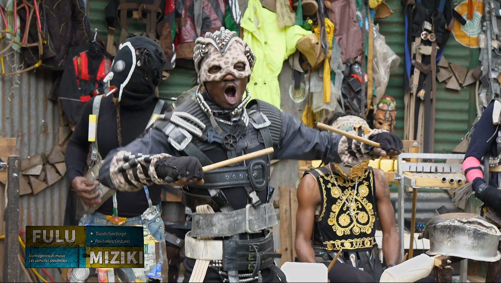 A performer in a pale mask and black costume raises two wooden beaters; other musicians and handmade instruments fill the background.