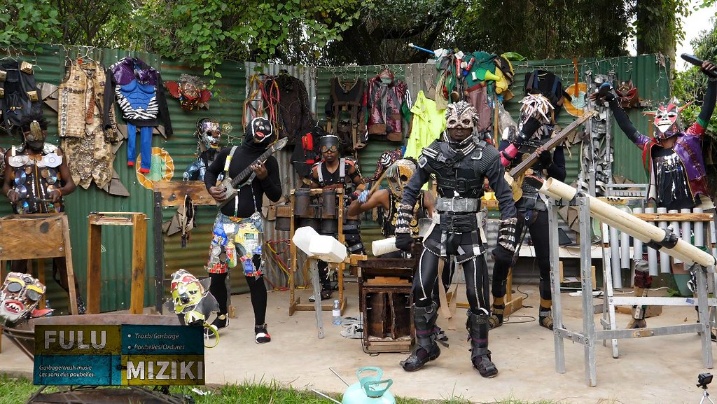 A group of masked musicians stands among instruments made with tubes, wooden frames and containers; costumes hang behind them.
