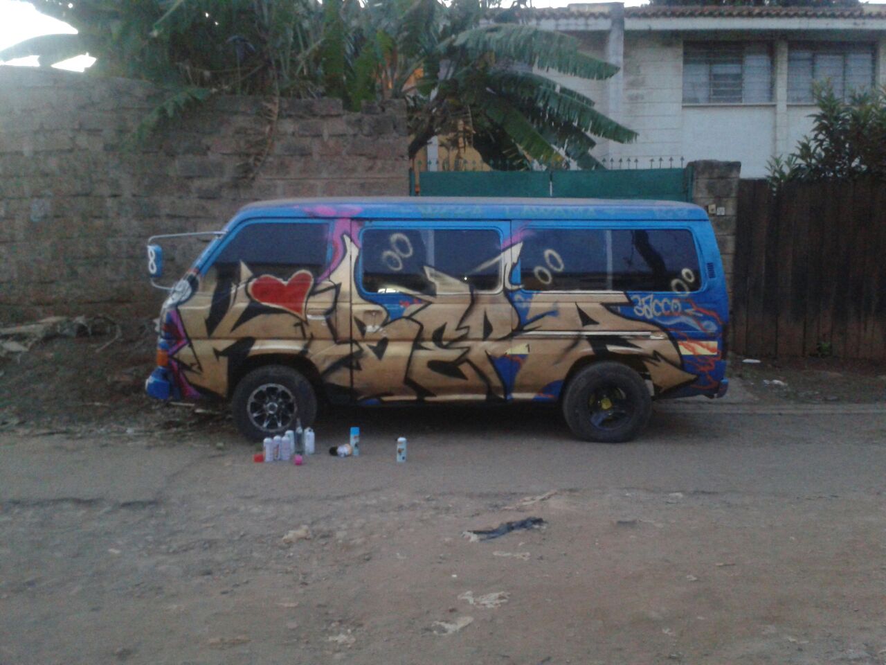 A blue minivan covered in large graffiti lettering, with paint containers nearby.