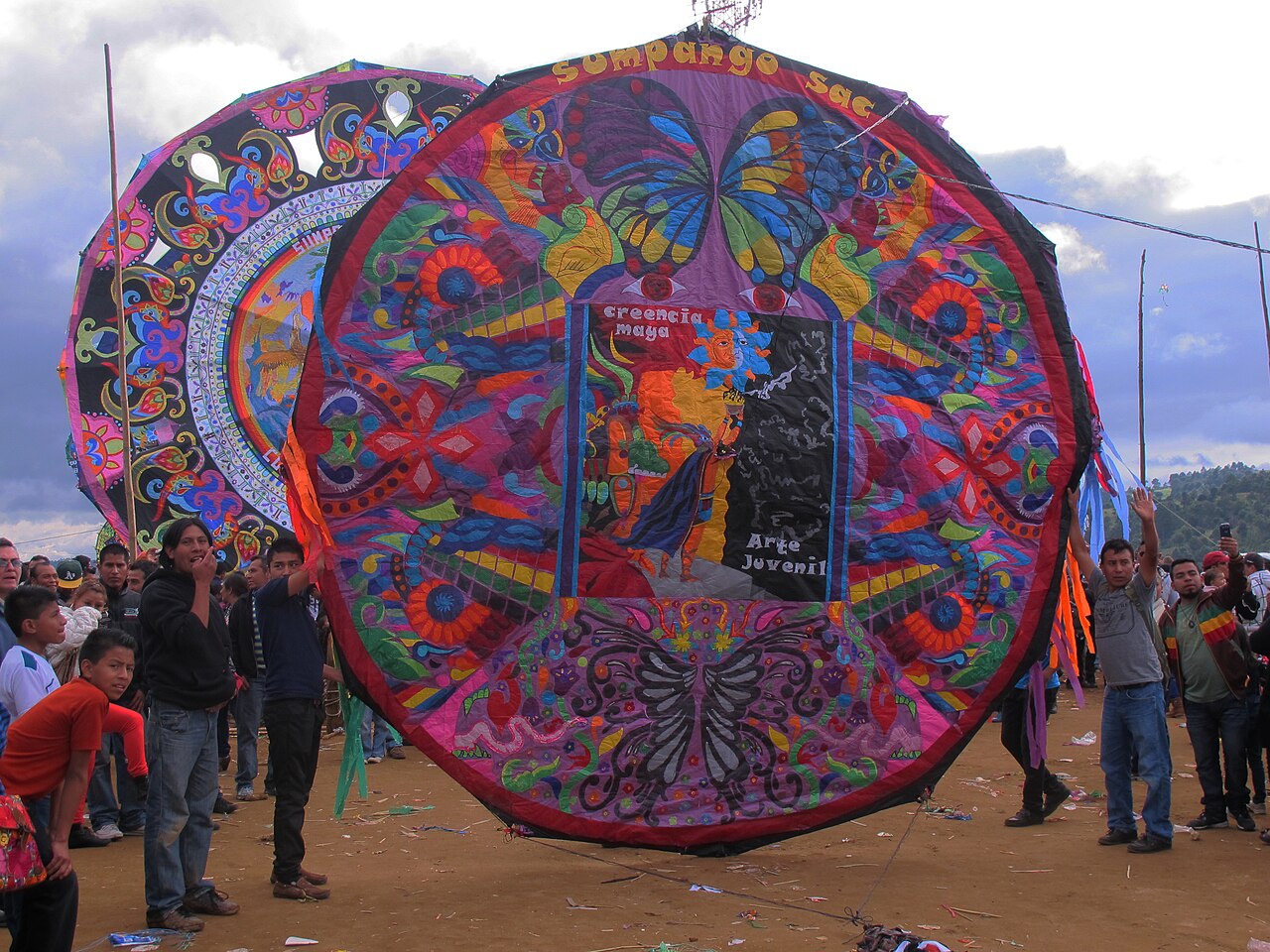 People hold a large circular kite decorated with butterflies and a central figure.