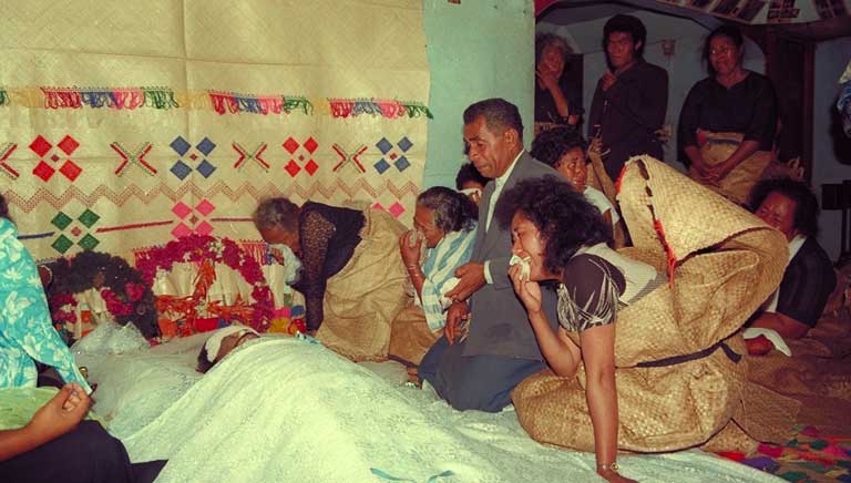 People wearing woven mats gather indoors beside flowers and a coffin.